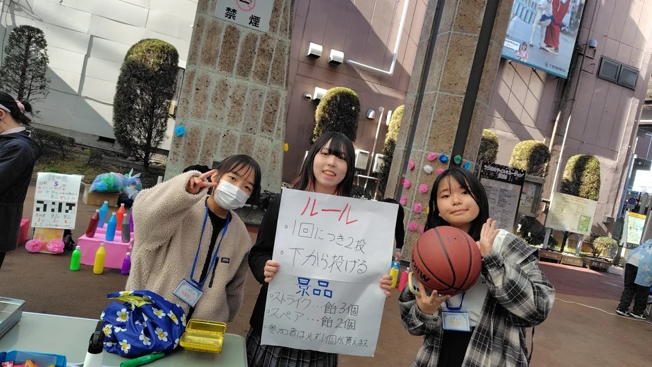 屋外のイベントで、3人の若い女性がゲームブースを運営しています。