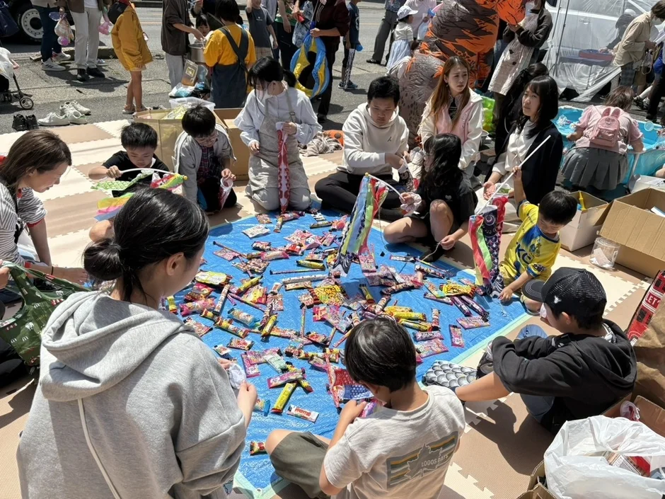 屋外のイベントで、多くの子供と大人が青いシートを囲んで座り、お菓子や鯉のぼり作りに興じている様子です。